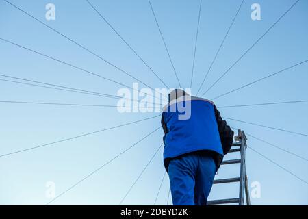 Elettricista ripara il cablaggio elettrico sul tetto di un alto edificio che si erge sulle scale contro il cielo blu. Spazio di copia Foto Stock