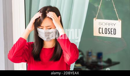 Asian giovane donna indossare maschera viso proteggere Sad avviso segno etichetta tavola di legno 'SORRY SIAMO chiusi SI PREGA DI TORNARE INDIETRO' appeso attraverso porta di vetro Foto Stock