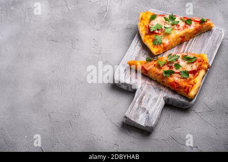 Fette di pizza calda con mozzarella, prosciutto, pomodoro e prezzemolo su tagliere in legno, fondo in pietra, angolo di visualizzazione per testo Foto Stock