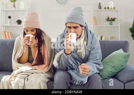 Giovane coppia malata avvolta in plaid seduti sul divano, bevendo tè caldo e cercando di riscaldare Foto Stock