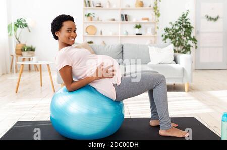 Esercizio durante il concetto di gravidanza. Madre nera positiva che lavora fuori sulla sfera di idoneità a casa Foto Stock