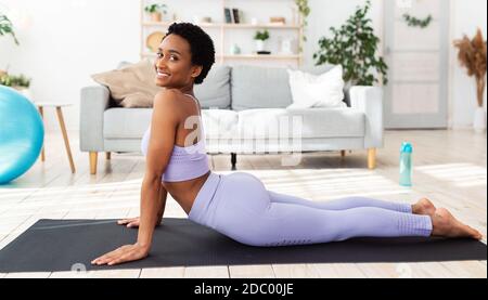 Calzare la giovane donna nera che pratica yoga sul tappeto sportivo a casa, rendendo la posa della cobra Foto Stock