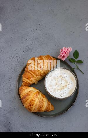 Colazione con tazza di caffè, croissant freschi e rosa su fondo di pietra grigia. Bella composizione. Concetto di caffè. Vista dall'alto, spazio di copia. Foto Stock