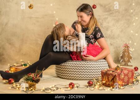 Scatto di studio di Natale di una bambina carina e di sua madre, una famiglia felice. Festivo, sfondo beige con regali, palle di Natale e luci Foto Stock