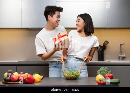 Ragazzo asiatico che dà regalo alla ragazza in cucina a casa Foto Stock