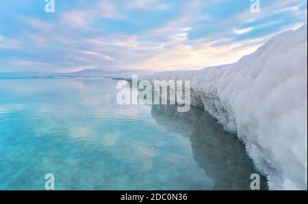 Sabbia completamente ricoperta di sale cristallino assomiglia a ghiaccio o neve sulla riva del Mar Morto, acque turchesi blu vicino, cielo colorato con dis del sole del mattino Foto Stock