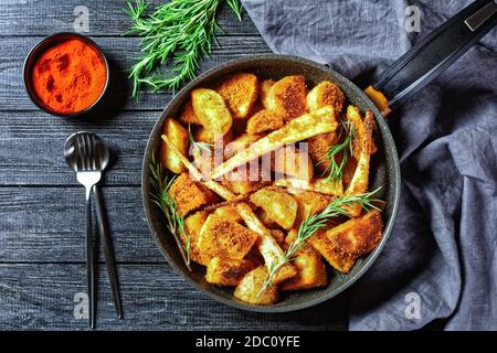 Verdure di radice al forno: Spicchi di patata e di pastinaca impanati con paprika affumicata e rametti di rosmarino fresco serviti su un backgrou di legno Foto Stock