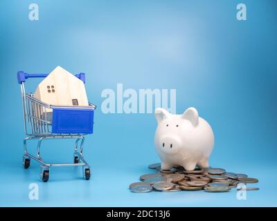 Una casa di legno modello su un carrello della spesa con un mucchio di monete e Piggy banca su uno sfondo blu. Concetto di ipoteca. Soldi e casa. Stato finanziario Foto Stock