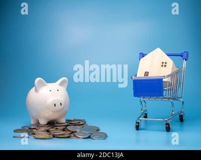 Una casa di legno modello su un carrello della spesa con un mucchio di monete e Piggy banca su uno sfondo blu. Concetto di ipoteca. Soldi e casa. Stato finanziario Foto Stock
