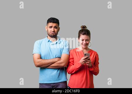 Ritratto di funny giovane uomo in abbigliamento casual in piedi con le braccia conserte, cercando risentita in telecamera, arrabbiato per la donna che utilizza il telefono e ignorando lui. è Foto Stock