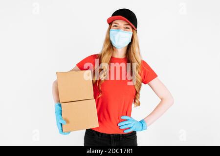 Un corriere in una uniforme rossa, indossando guanti di gomma e una maschera medica tiene scatole di cartone isolate su uno sfondo bianco. Un dipendente femminile lavora come Foto Stock