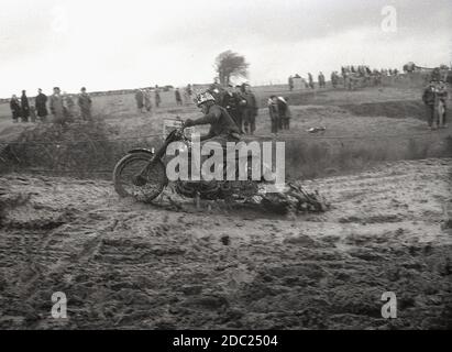 Alla fine degli anni '50, un concorrente maschile in un evento di scrambling motociclistico, che attraversa un sentiero fangoso. Inventata nel 1924, a Camberley, Surrey, in Inghilterra, le moto usate per la corsa negli anni '50 e all'inizio degli anni '60 erano poco diverse dalle moto da strada del tempo, con pochissime sospensioni. Oggi lo sport è conosciuto come motocross – il nome francese per il cross-country motociclismo – e chiamato Supercross negli Stati Uniti. Foto Stock