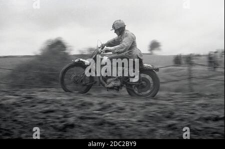 Alla fine degli anni '50, un concorrente maschile in un evento di scrambling motociclistico, che attraversa un sentiero fangoso. Inventata nel 1924, a Camberley, Surrey, in Inghilterra, le moto usate per la corsa negli anni '50 e all'inizio degli anni '60 erano poco diverse dalle moto da strada del tempo, con pochissime sospensioni. Oggi lo sport è conosciuto come motocross – il nome francese per il cross-country motociclismo – e chiamato Supercross negli Stati Uniti. Foto Stock