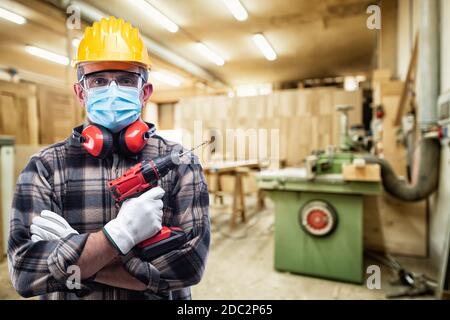 Falegname in officina, indossa casco, occhiali, guanti in pelle, cuffie e maschera chirurgica per prevenire l'infezione da coronavirus. Preven Foto Stock