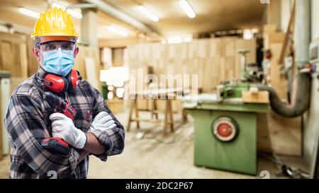 Falegname in officina, indossa casco, occhiali, guanti in pelle, cuffie e maschera chirurgica per prevenire l'infezione da coronavirus. Preven Foto Stock