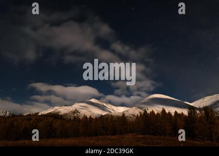 Paesaggio notturno panoramico con nuvole e stelle nel cielo su montagne innevate e foresta al chiaro di luna in autunno Foto Stock