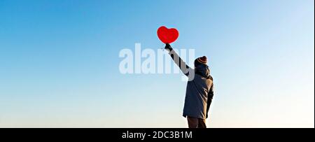 giovane uomo dalla schiena in abiti caldi che tiene un palloncino rosso a forma di cuore contro il cielo blu. San Valentino e romantico concetto, copia spazio Foto Stock