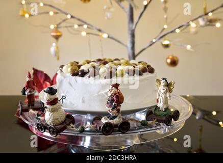 Torta di Natale ghiacciata su un supporto trasparente con noci di cioccolato sulla parte superiore, figure di Natale intorno alla base e un albero di Natale illuminato in ramoscello sullo sfondo Foto Stock