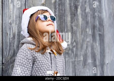 Bambina cool che indossa cappello Babbo Natale e occhiali da sole in piedi Contro uno sfondo grunge in legno all'aperto nel tempo di Natale - Buon Natale e nuovo Yea Foto Stock