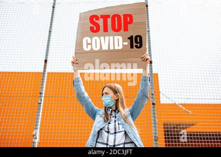 Una giovane donna in una maschera medica protettiva tiene un cartello di cartone lettura Stop il coronavirus, in piedi in città contro una parete grigia. Quarantin Foto Stock