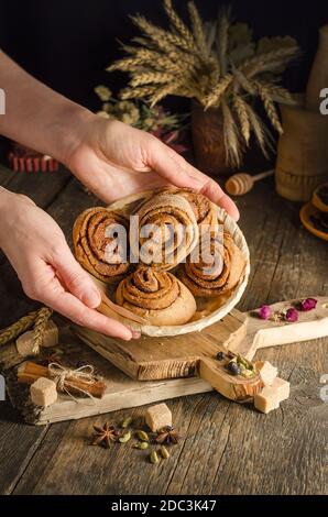 Donna che serve focaccine di cannella fatte in casa in un piatto di vimini su uno sfondo scuro, foto di cibo di stile scuro Foto Stock