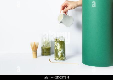 Donna mano versando il latte in vetro con tè matta verde ghiaccio su sfondo bianco. Latte opaco ghiacciato. Foto Stock