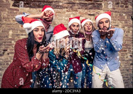 Buon giorno di Natale - sorridendo la gente con la maschera di faccia giù Soffiare i confetti dalle mani - amici che festeggiano all'interno del Serata nel tempo del coronavirus - nuovo No Foto Stock