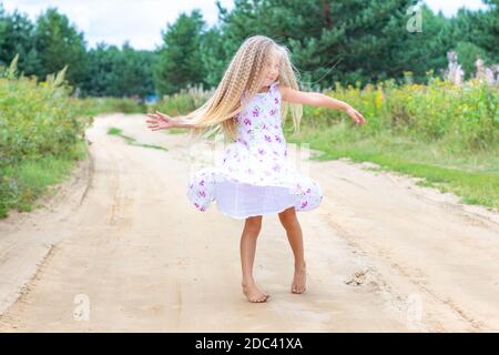 Una ragazza con lunghi capelli ricci biondi sta ballando, girando su una strada forestale. Infanzia, felicità, libertà di movimento, danza, riunificazione con la natura. Foto Stock