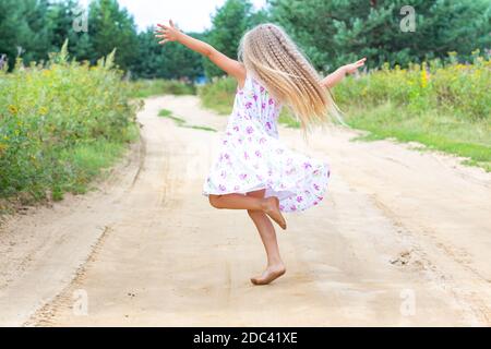 Una ragazza con lunghi capelli ricci biondi sta ballando, girando su una strada forestale. Infanzia, felicità, libertà di movimento, danza, riunificazione con la natura. Foto Stock