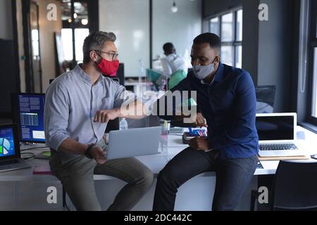 Diversi colleghi maschili che indossano maschere viso si salutano l'un l'altro toccare i gomiti mentre in moderno di Foto Stock