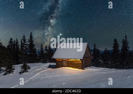 Fantastico paesaggio invernale con casa in legno in montagne innevate. Cielo stellato con Via Lattea e capanna innevata. Vacanza di Natale e concetto di vacanza invernale Foto Stock