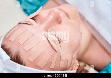 Kinesiotaping. Giovane donna sdraiata con nastro sul viso. Fisioterapia e procedura cosmetologica. Metodo di ringiovanimento non chirurgico della pelle. Pelle del viso Foto Stock