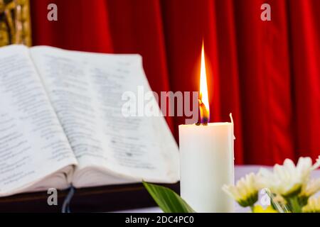 la bibbia si apre su un tavolo con candela e tenda rossa come sfondo. fiore come primo piano. Perfetto per la religione, pasqua e natale temi. Candela fucus Foto Stock