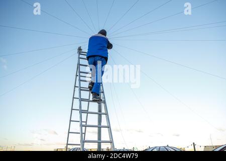 Elettricista ripara il cablaggio elettrico sul tetto di un alto edificio che si erge sulle scale contro il cielo blu. Spazio di copia Foto Stock