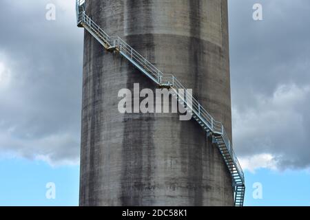 Grande silo industriale cilindrico in calcestruzzo con scala in acciaio che si avvolge sotto nuvola di tempesta scura. Foto Stock