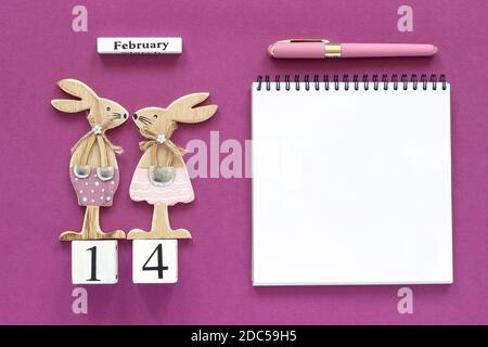 Composizione romantica cubi calendario 14 febbraio Coppia di conigli figurine in legno amanti del legno, taccuino vuoto aperto per testo con penna su sfondo viola C Foto Stock