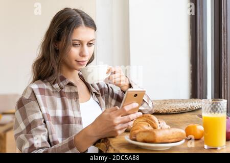 Donna che guarda i social network utilizzando il cellulare, gustando una sana colazione. Foto Stock