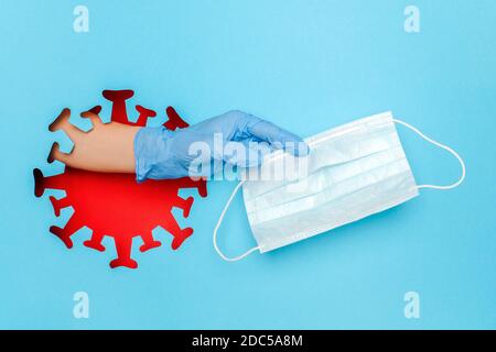 Mano in guanto di lattice dà maschera medica attraverso il foro in forma di coronavirus rosso, virus, batteri, microbi su sfondo blu. Concetto creativo di prote Foto Stock