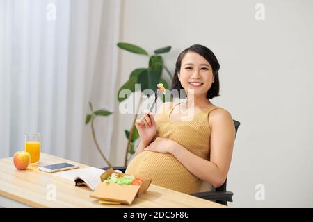 donna incinta felice che mangia insalata mentre si siede in ufficio vicino tabella Foto Stock