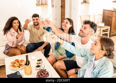Gruppo di amici di corsa misti che tostano con champagne festeggiando a. home - giovani multirazziali seduti sul divano che si aggraffa bicchieri di vino bianco Foto Stock