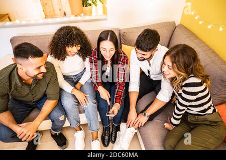 Gruppo di amici di corsa misti che si divertono a casa seduta Sul divano guardando lo smartphone - multirazziale giovane persone che utilizzano il social network per la condivisione Foto Stock
