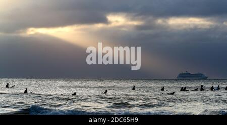 Bournemouth, Regno Unito. 19 novembre 2020. In primo piano. Un pozzo di luce solare illumina una delle navi da crociera ormeggiate a Poole Bay mentre i surfisti aspettano di prendere un'onda all'alba sulla spiaggia di Bournemouth sulla costa del Dorset. Credit: Richard Crease/Alamy Live News Foto Stock