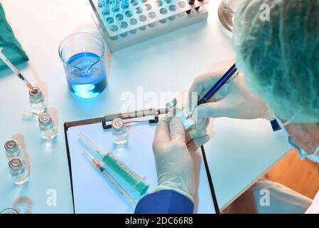 Operatore medico dotato di protezione personale scrittura su fiala su tavolo da laboratorio. Vista dall'alto. Composizione orizzontale. Foto Stock