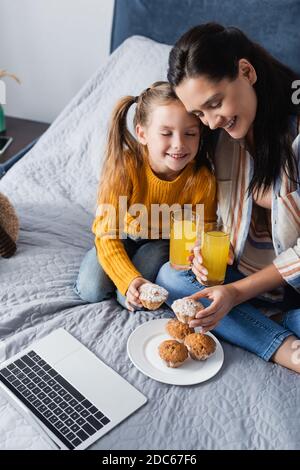 madre e figlia felici che tengono il muffin e il succo d'arancia vicino computer portatile Foto Stock