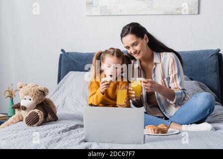 un bambino entusiasta con una madre felice che guarda il film su un computer portatile con succo d'arancia e muffin Foto Stock