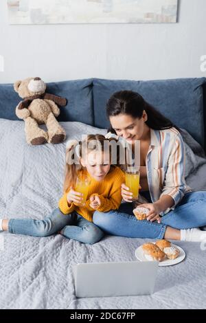 vista ad alto angolo di una donna sorridente e di un'avvincente figlia film su computer portatile con muffin e succo d'arancia Foto Stock