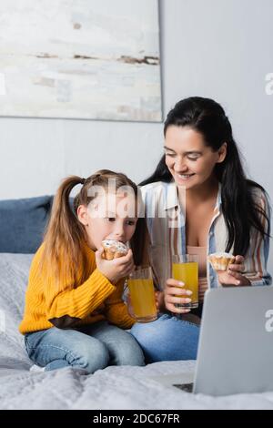 madre felice che guarda la figlia che mangia il muffin e guarda il film su un computer portatile Foto Stock