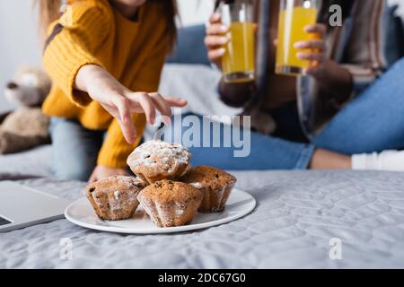 vista ritagliata del bambino che prende il muffin dal piatto vicino alla mamma tenendo il succo d'arancia su sfondo sfocato Foto Stock