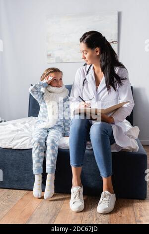 medico che scrive nella scheda ambulatoriale mentre sedendo vicino alla ragazza malata indossare pigiami Foto Stock