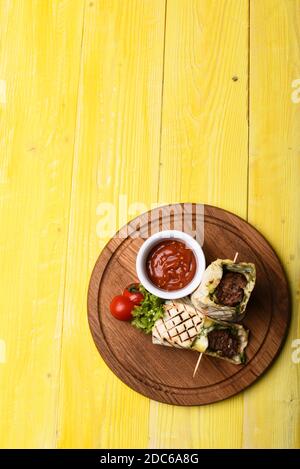 Concetto di portata principale. Carne macinata avvolta con pane di lavasina su fondo giallo di legno. Flatbread con ripieno di maiale o pollo e salsa di peperoncino rosso. Burrito o tortilla serviti in ristorante o caffetteria. Foto Stock